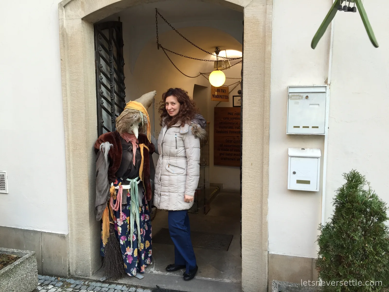 Inga standing beside a life-size witch marionette in the arched doorway of a Prague puppet shop with a Loutky Marionettes sign