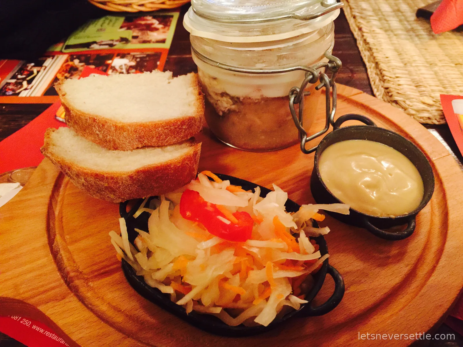A Czech pub starter on a wooden board: a preserving jar of pate under a layer of fat, bread slices, pickled cabbage slaw and mustard