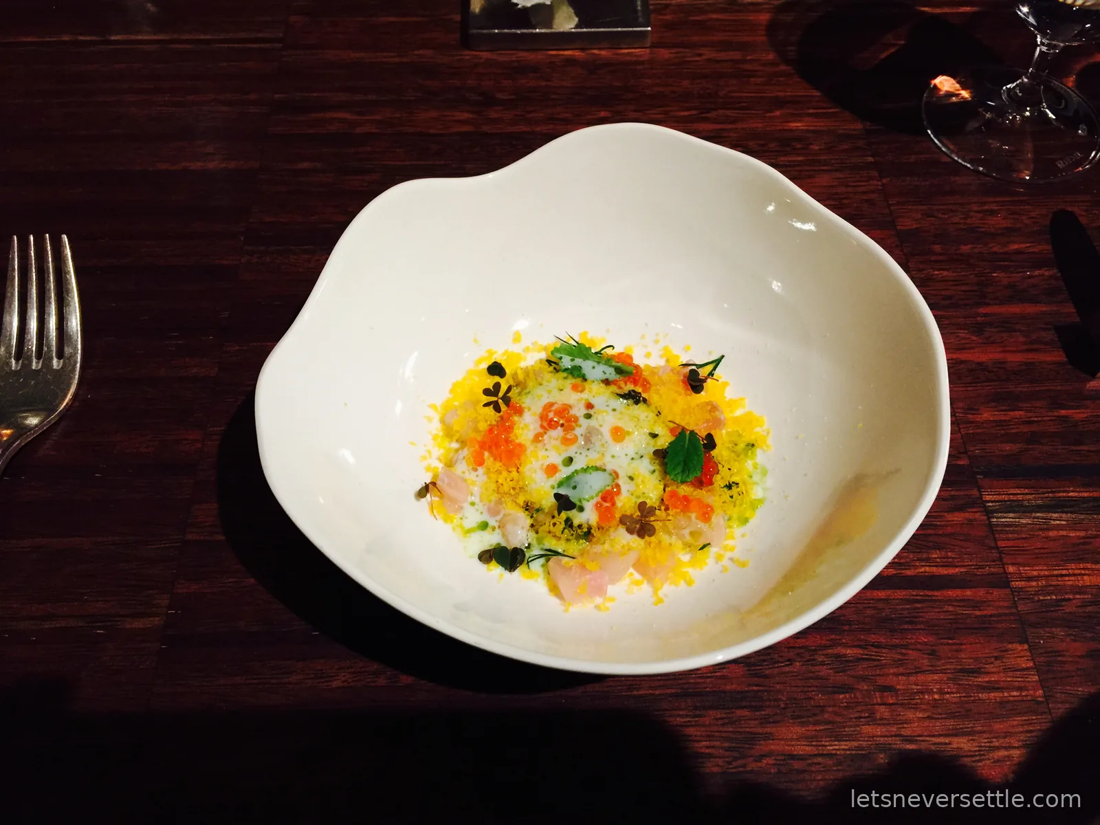 A sculpted white bowl holding a fine dining course scattered with orange roe, golden crumb and tiny herb leaves