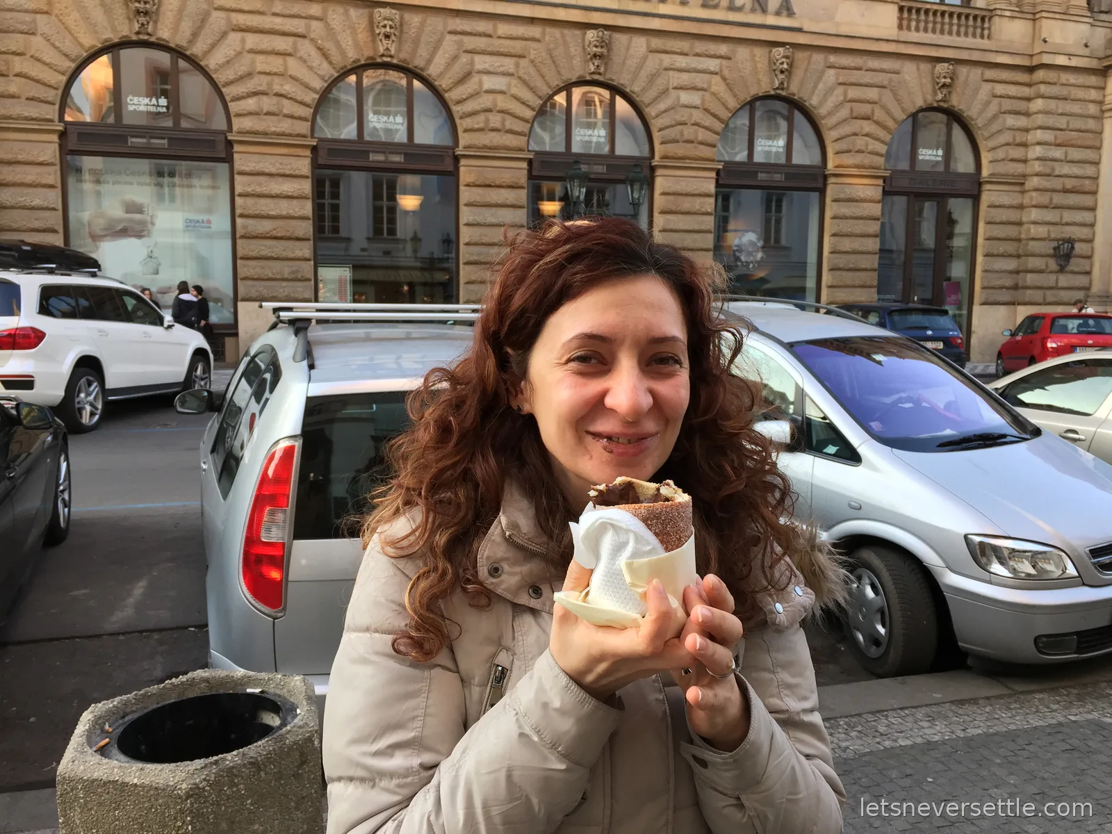Inga smiling with a chocolate-filled trdelnik chimney cake wrapped in a napkin on a Prague street, arched stone facades behind