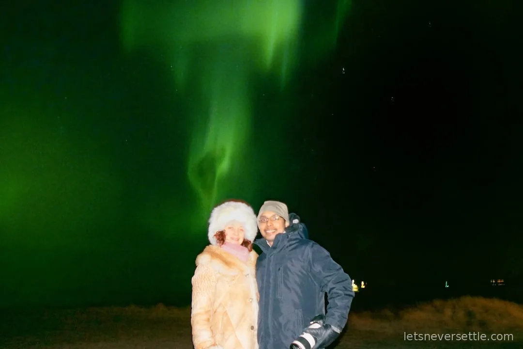 Inga and Dileep standing together under a bright green aurora filling the night sky