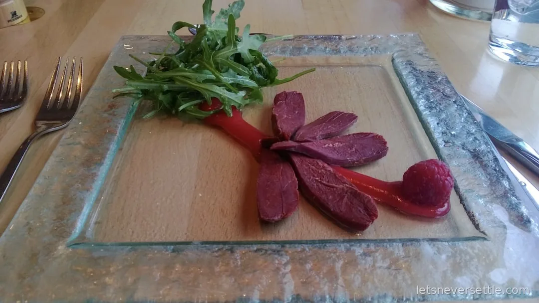 Slices of dark cured meat fanned on a glass plate with rocket and a raspberry