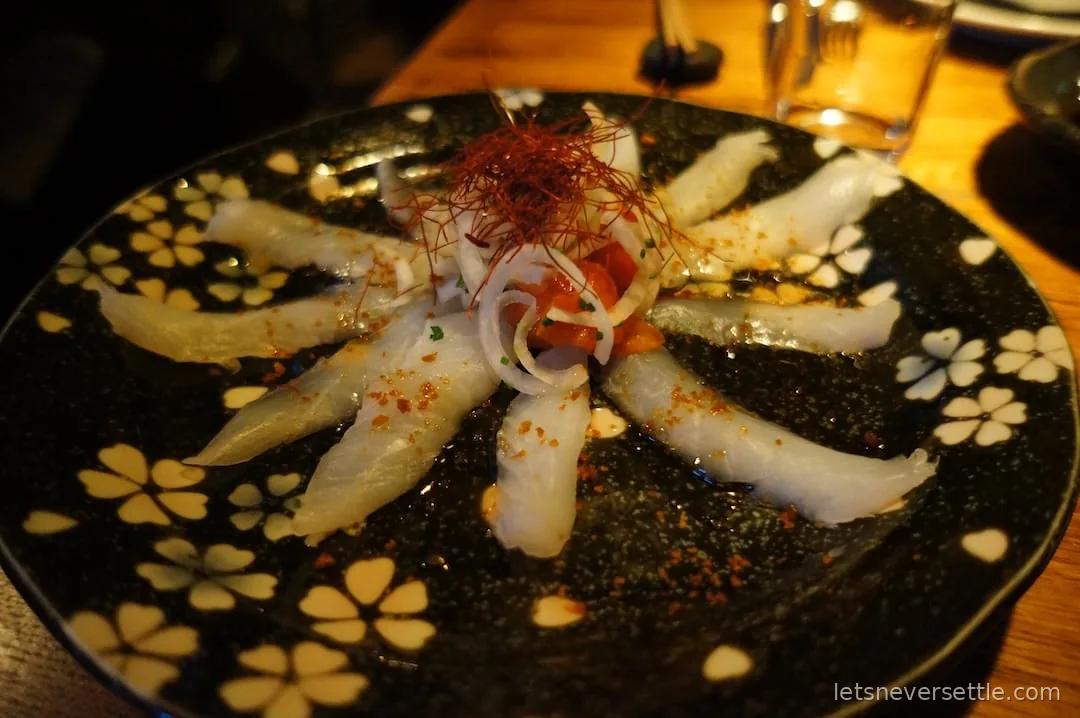 Thin slices of white fish carpaccio arranged on a black plate patterned with pale flowers
