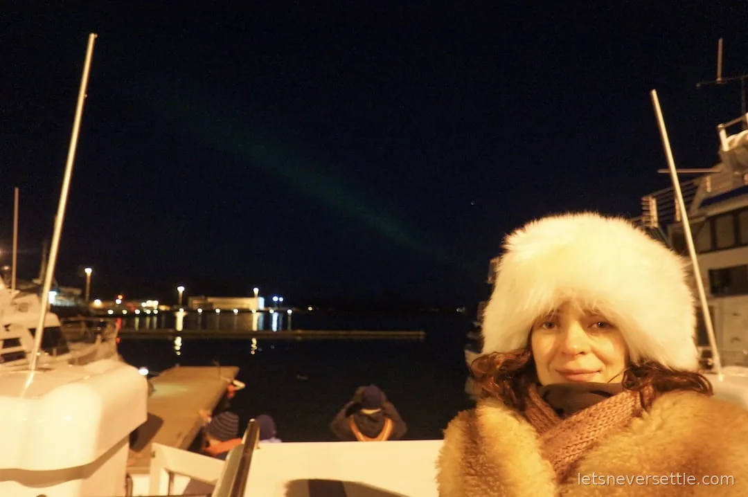 Inga in a white fur hat on a boat in Reykjavik old harbour at night, a faint green aurora over the water behind