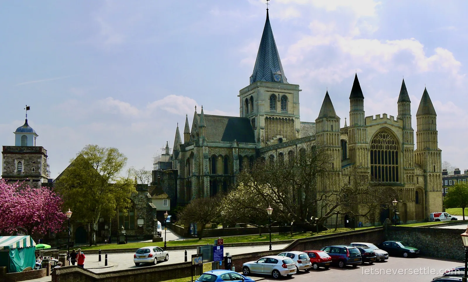 Rochester Cathedral with its grey spire and pinnacled west front, seen across a car park with pink blossom on the left