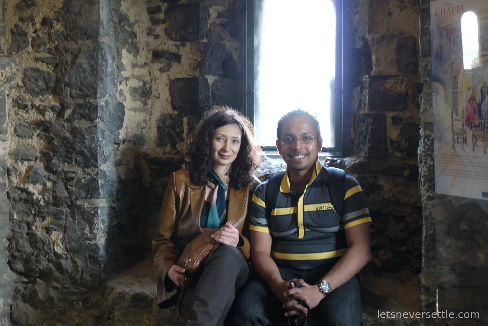Inga and Dileep sitting in a deep stone window recess of Rochester Castle, next to an information panel about the chapel