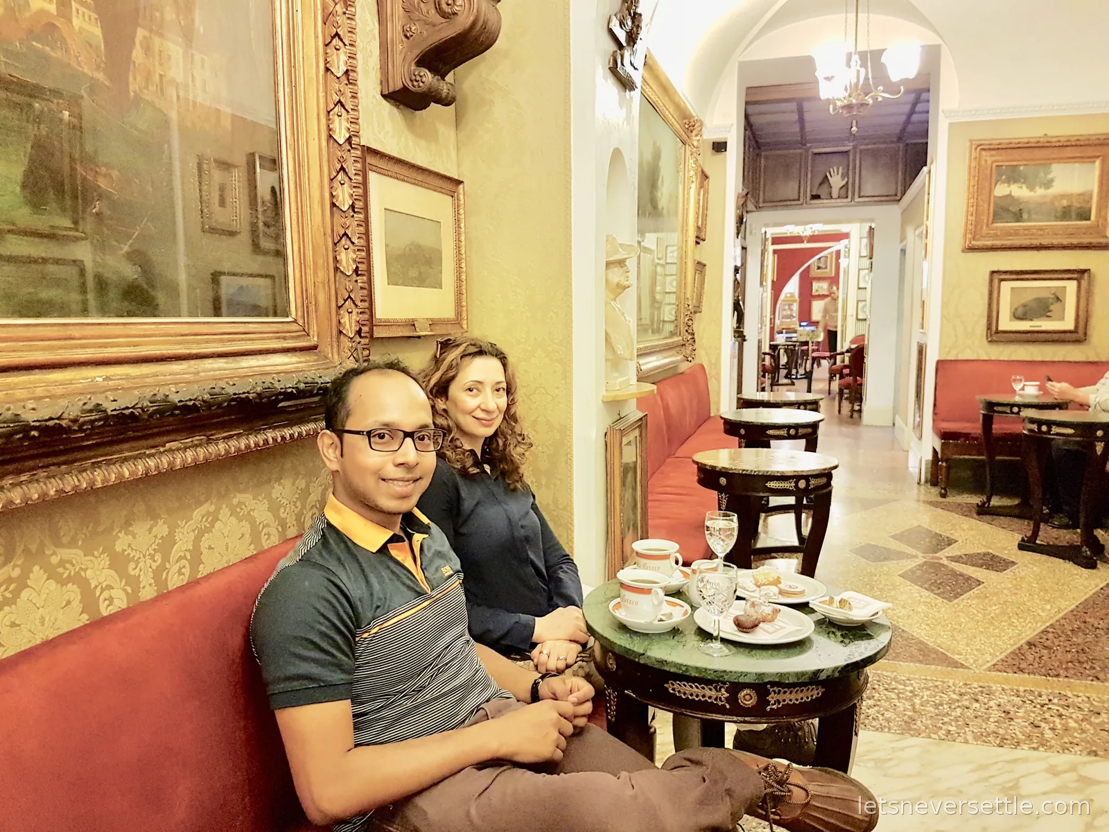 Inga and Dileep on a red velvet banquette in an old cafe with gilt framed paintings covering the walls and espresso cups on a marble table
