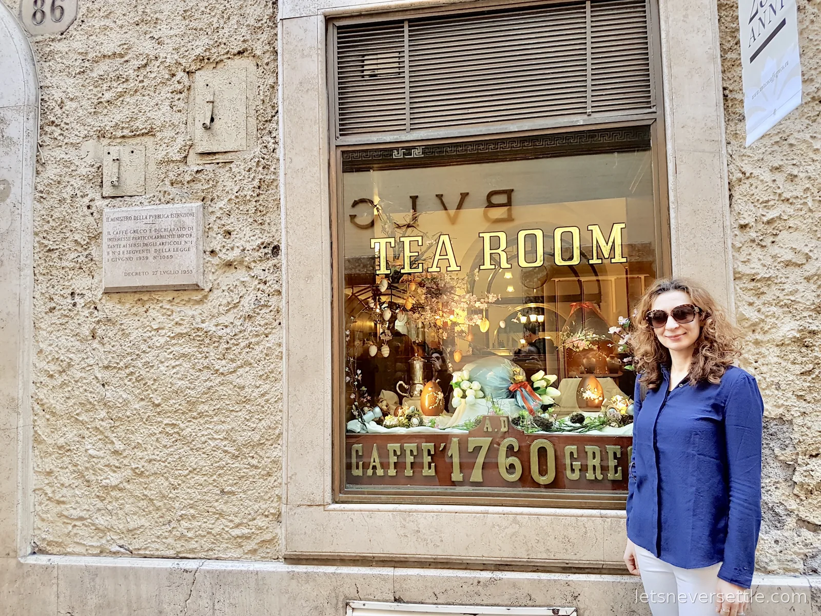 Inga standing beside a cafe window lettered Tea Room and Caffe 1760, with a chandelier and a display of pastries inside