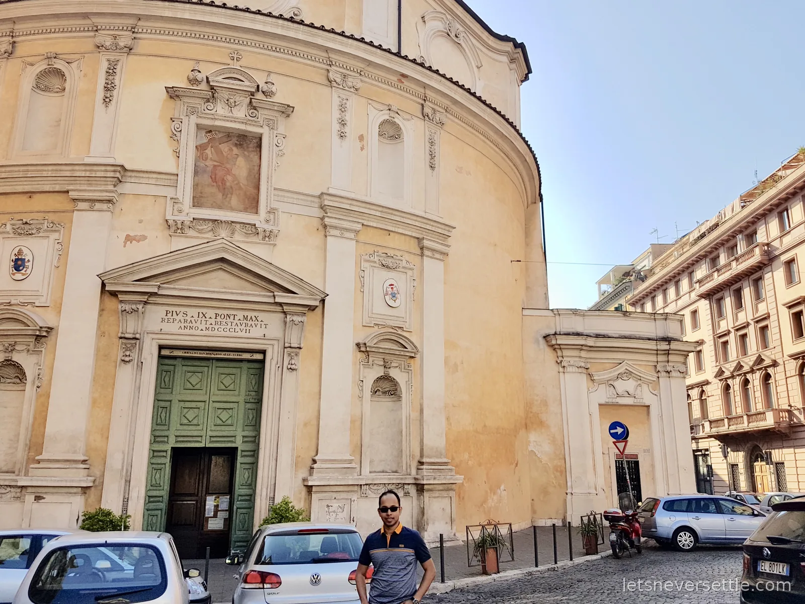 Dileep standing outside a curved pale yellow church front with a Latin inscription and green doors, cars parked in front
