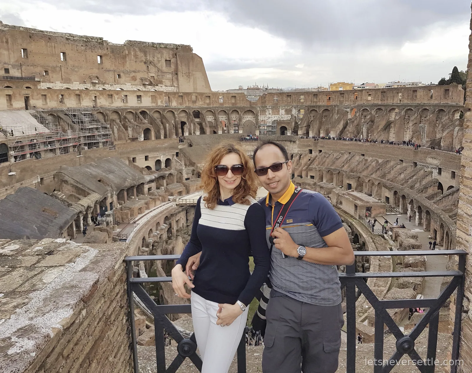 Inga and Dileep standing at a railing inside the Colosseum with the tiers of seating and the exposed underground chambers behind them