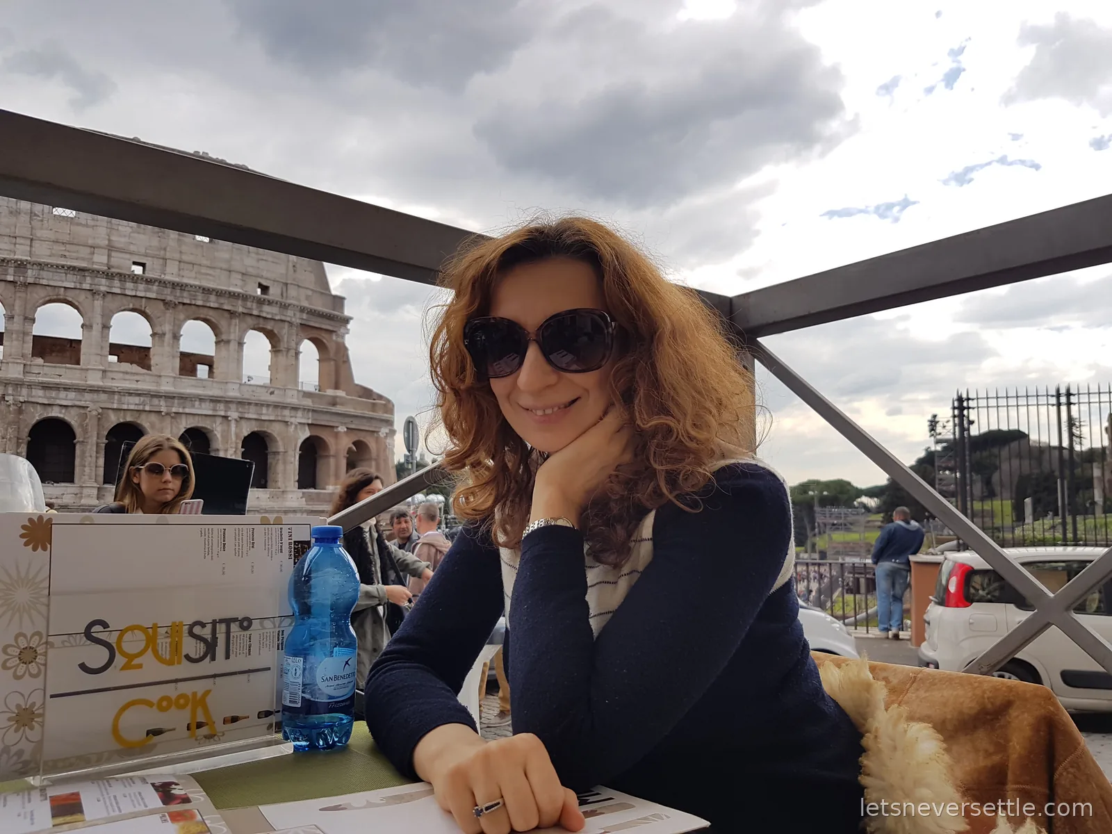 Inga in sunglasses leaning on a cafe table under an awning with the arches of the Colosseum behind her