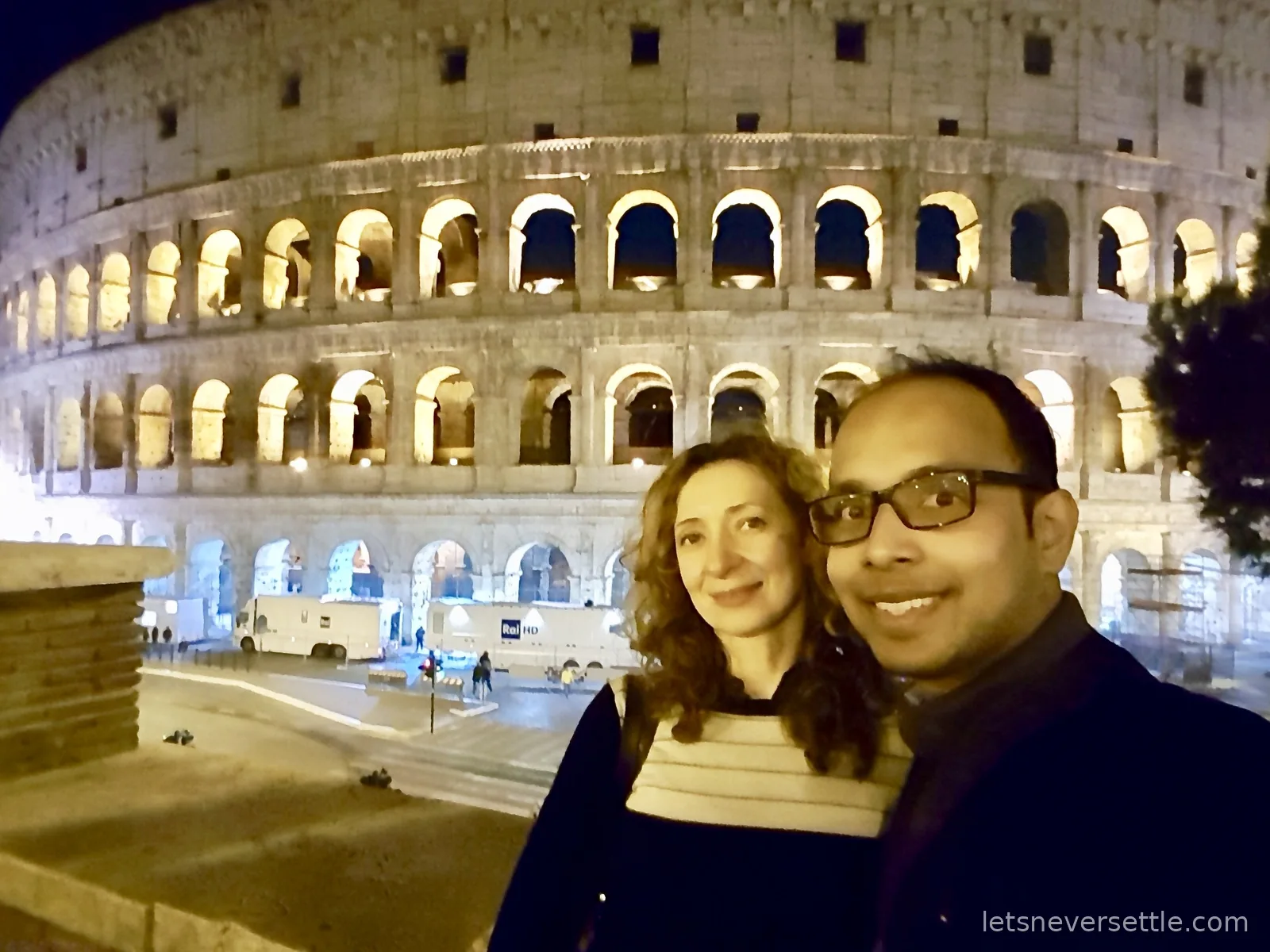 Inga and Dileep taking a selfie at night with the floodlit Colosseum behind them