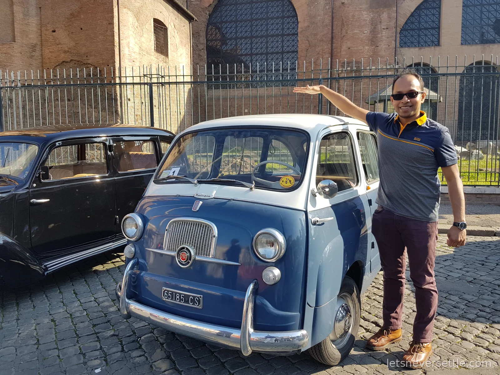 Dileep resting a hand on the roof of a small blue and white Fiat 600 Multipla parked beside a black vintage saloon on cobbles