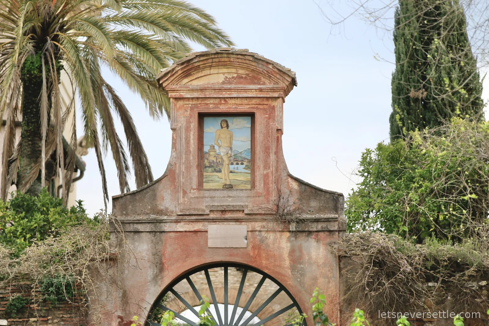A weathered pink stucco gateway topped with a painted tile of Saint Sebastian, framed by a palm and a cypress