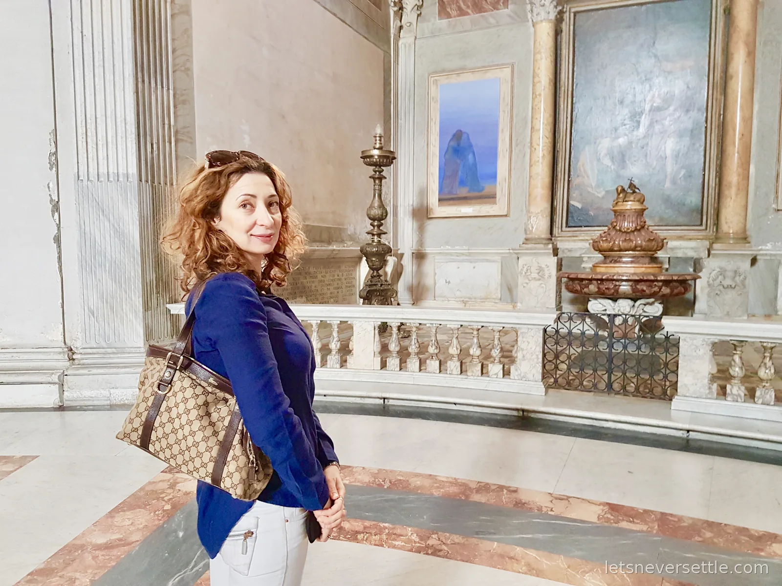 Inga looking over her shoulder in a marble gallery hung with old paintings, with a stone balustrade and gilt urns behind her