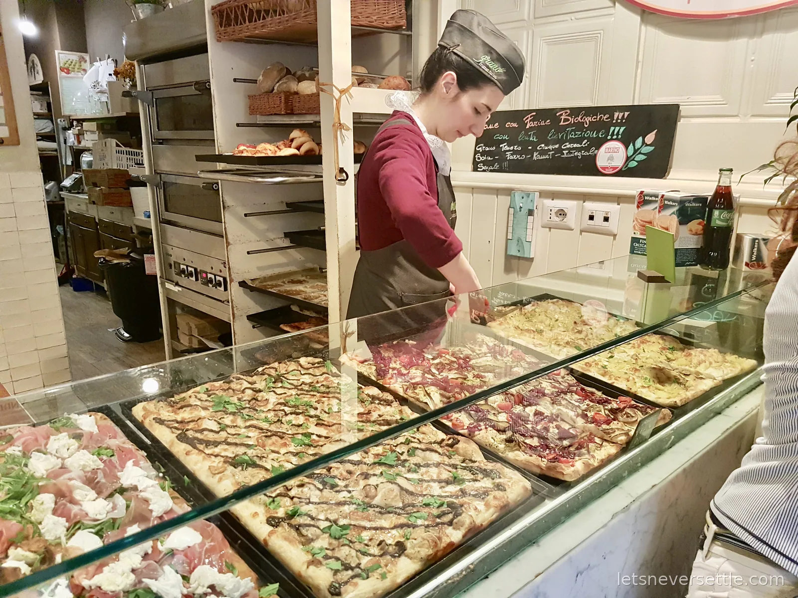 A woman in an apron cutting a tray of pizza behind a glass counter stacked with rectangular pizzas and a bread oven behind