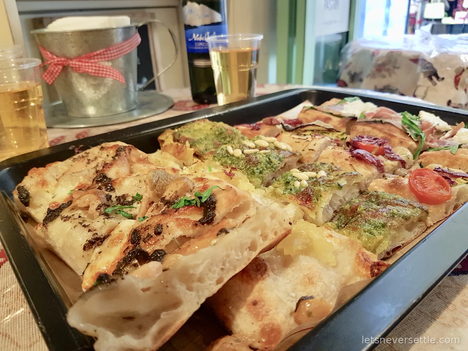 A metal tray of assorted pizza slices with toppings including tomato, rocket, pesto and cheese, with two plastic cups of wine beside it