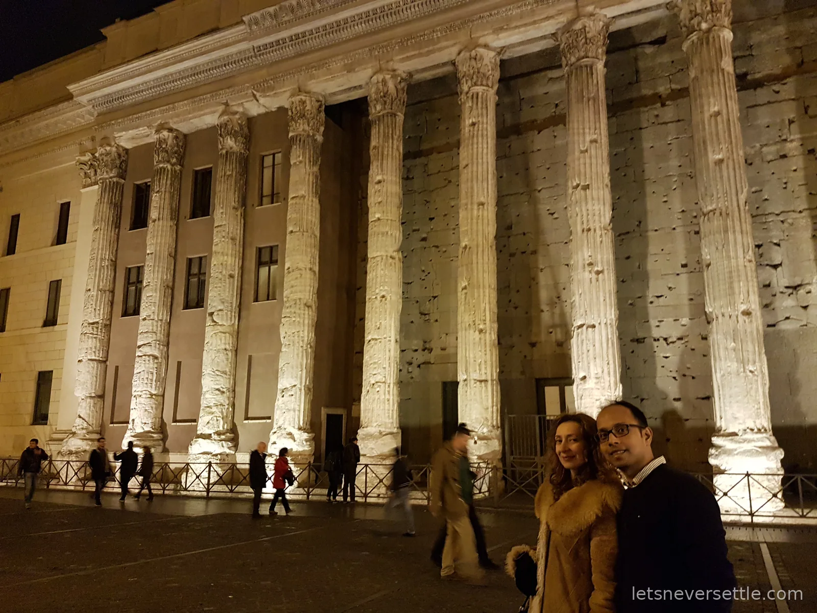 A row of enormous fluted Roman columns floodlit at night, built into the front of a later building, with people walking past