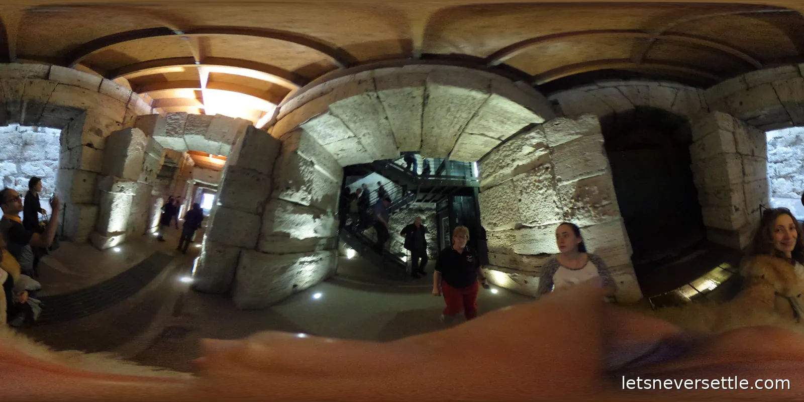 A vaulted stone passageway lit from below, with a steel staircase and visitors walking through