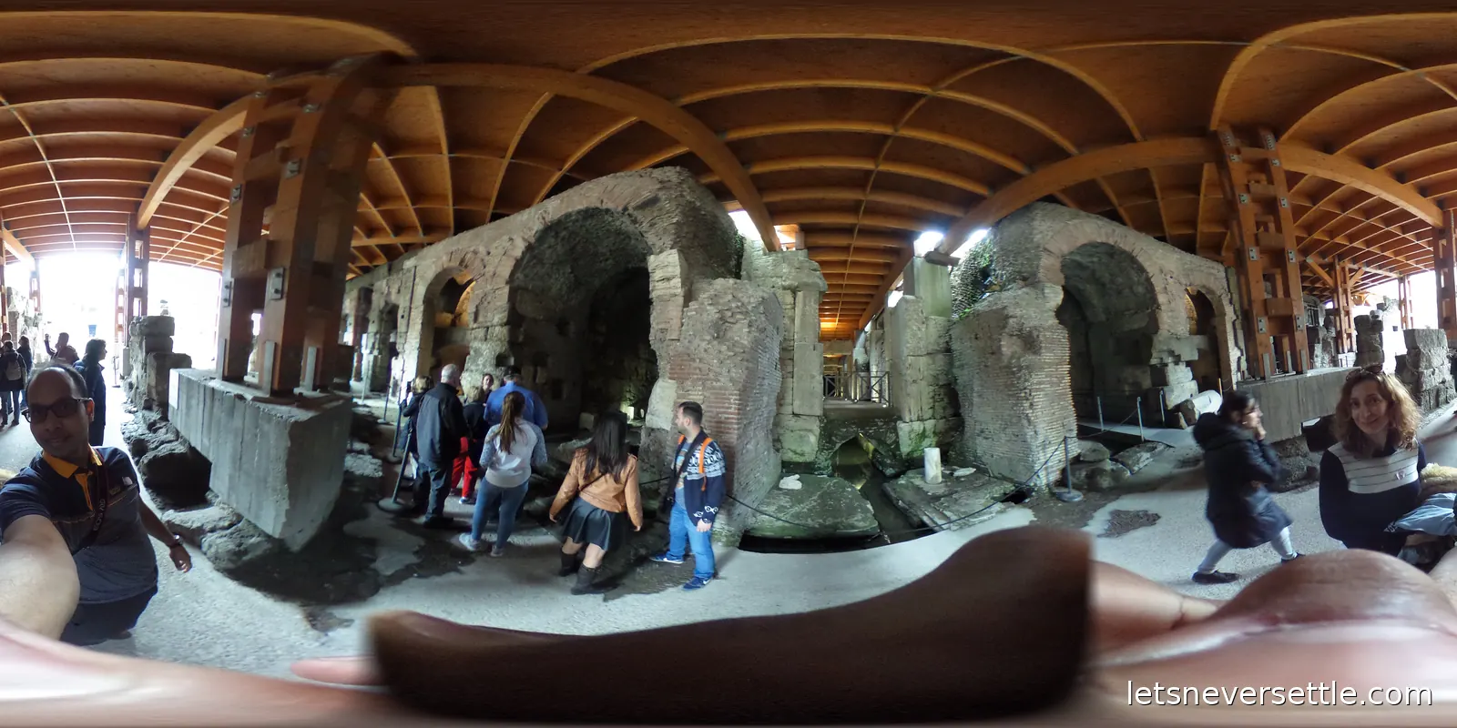 Brick Roman ruins sheltered under a curved timber roof, with visitors walking between the walls