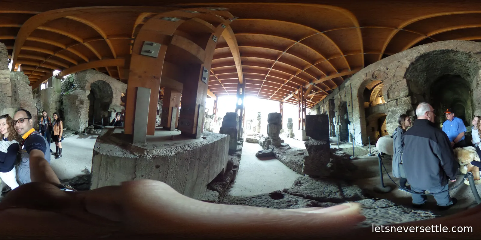 Carved stone fragments standing on concrete plinths among excavated brick walls under a wooden vaulted shelter