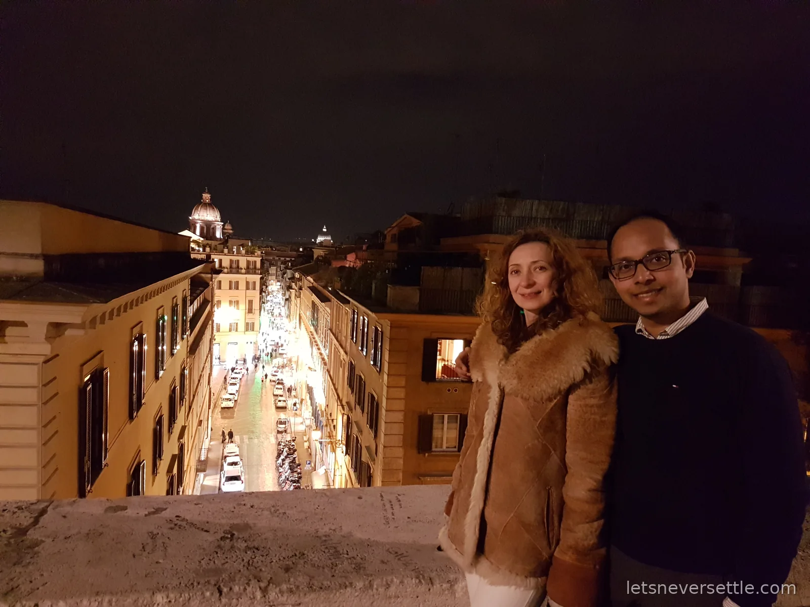 Inga and Dileep on a terrace at night above a lit shopping street running away between tall buildings, with domes in the distance