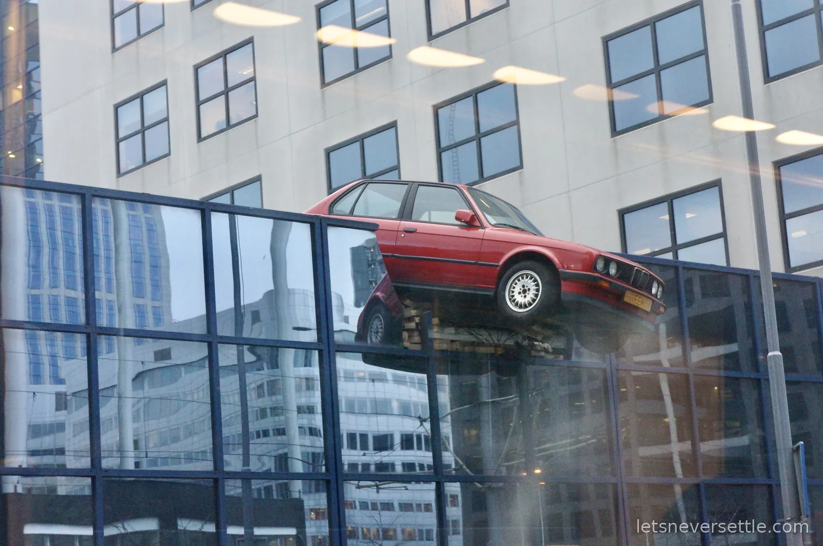 A red BMW perched on top of a glass gallery wall in front of an office block in central Rotterdam