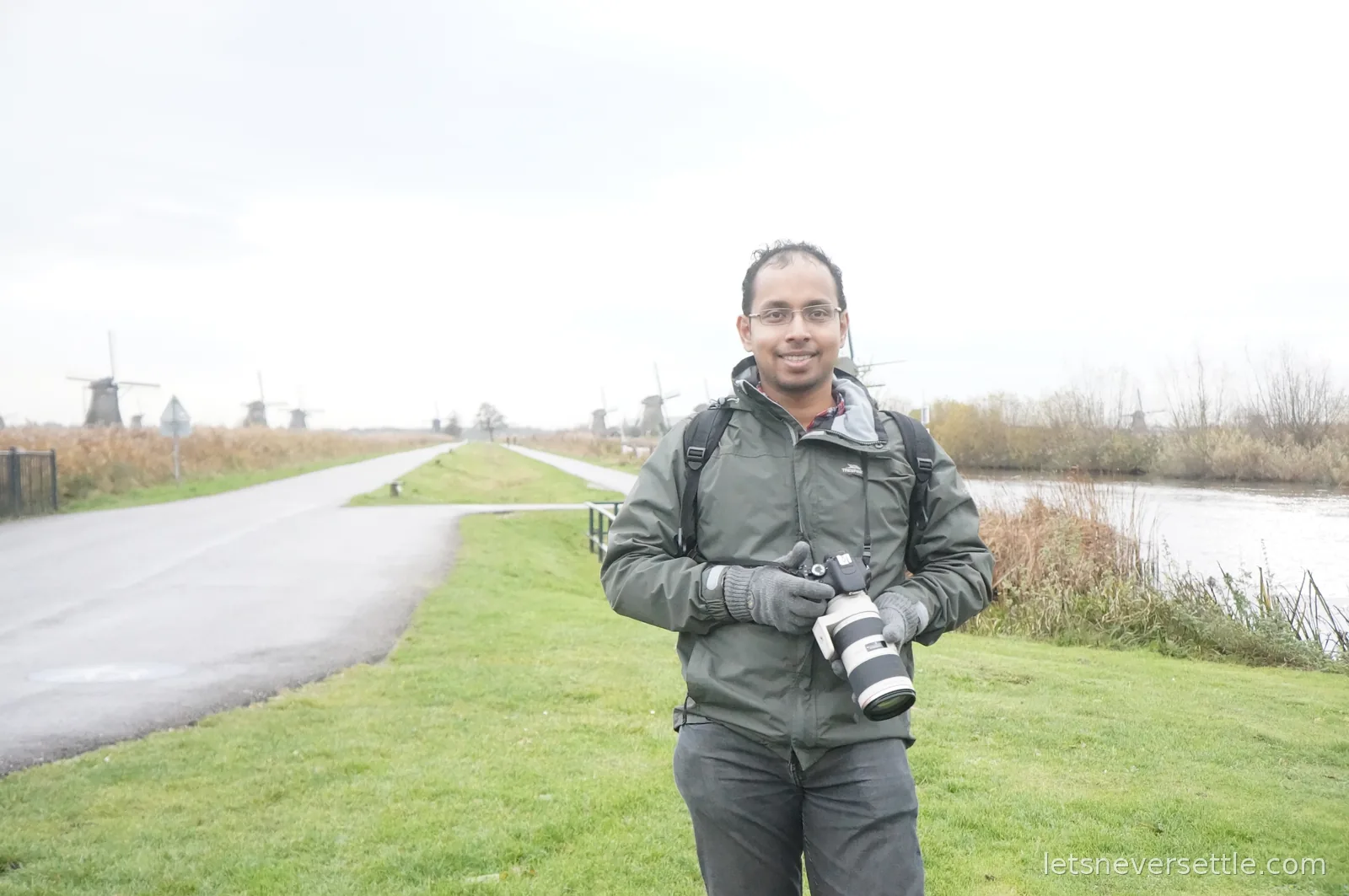 Dileep in a grey rain jacket holding a Canon camera with a white telephoto lens, a line of Kinderdijk windmills fading into the distance behind him