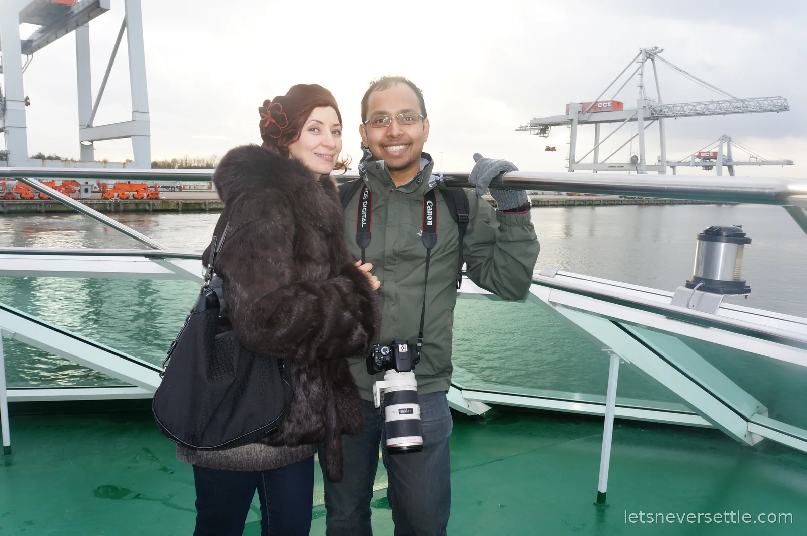 Inga and Dileep on the open deck of a harbour tour boat in Rotterdam, giant container cranes across the water behind them