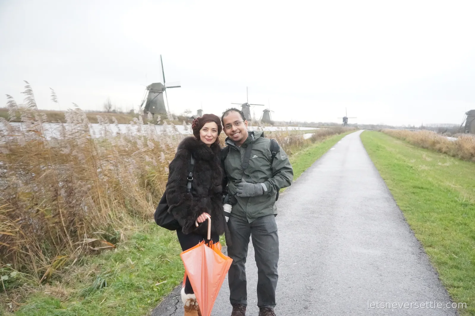 Inga and Dileep standing together on a dike path at Kinderdijk with windmills and reed beds stretching behind them