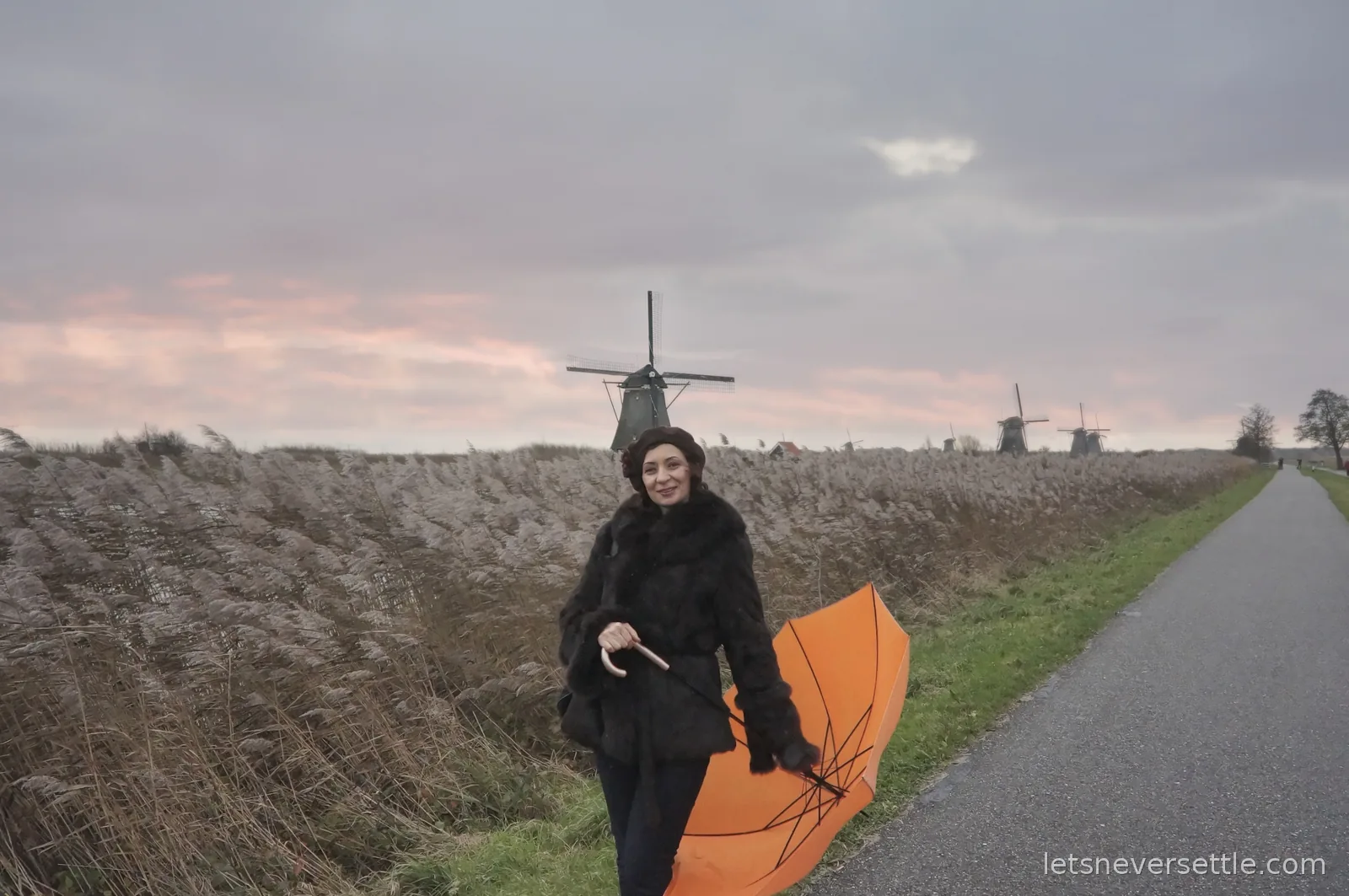 Inga in a fur coat and beret on a path at Kinderdijk holding an orange umbrella turned inside out, windmills on the skyline behind her