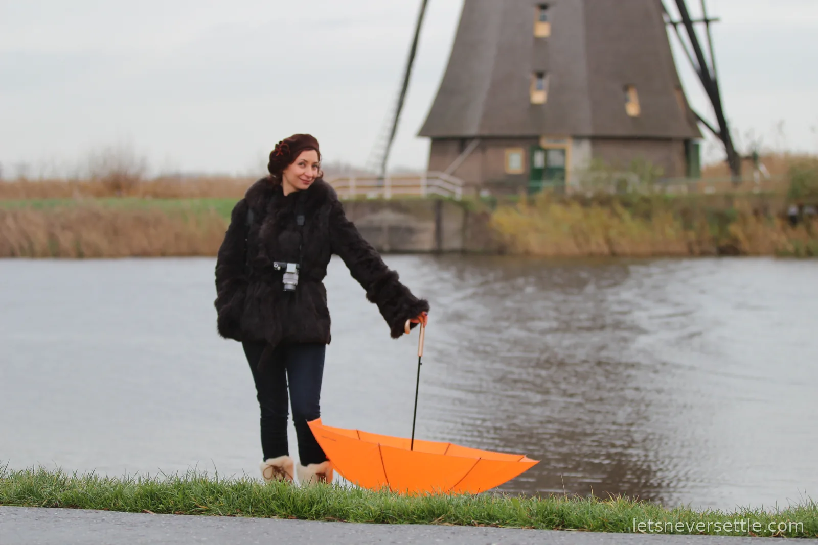 Inga standing at the edge of a canal at Kinderdijk trailing an upside-down orange umbrella, a thatched windmill on the far bank