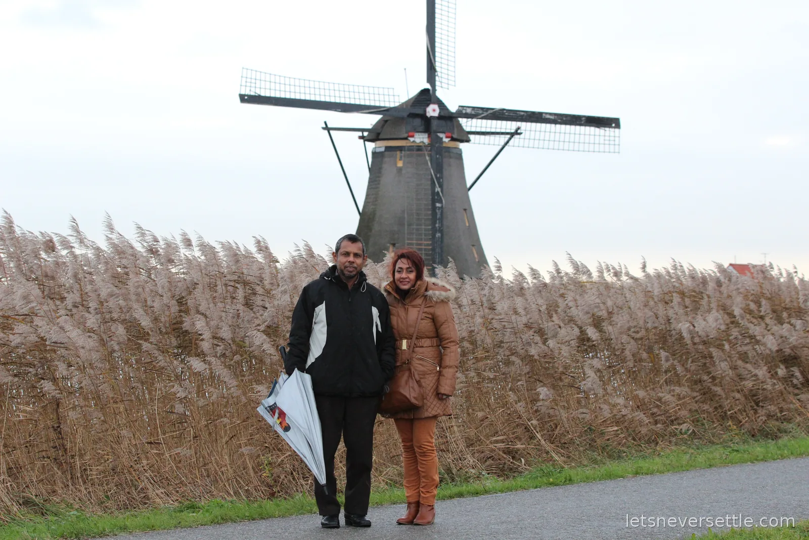 Dileep's parents standing side by side on a path in front of a large thatched windmill and wind-blown reeds at Kinderdijk