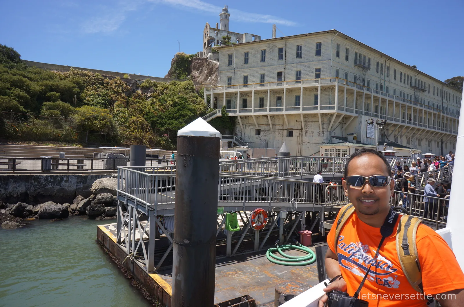 Dileep at the Alcatraz ferry dock with the prison buildings and lighthouse on the rock above