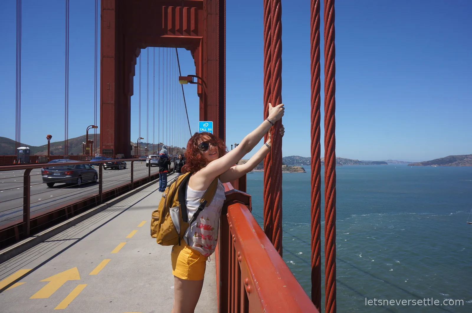 Inga reaching up to hold the suspension cables on the Golden Gate Bridge walkway, with traffic alongside and the bay far below