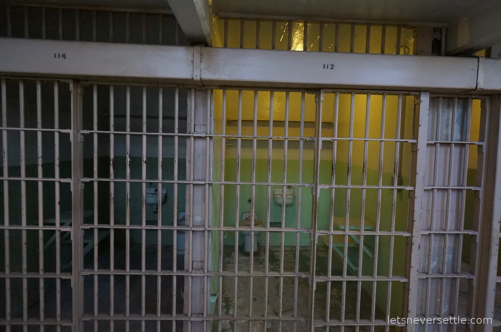 Two open prison cells with green walls, numbered 112 and 114, behind cream painted bars in the Alcatraz cell house