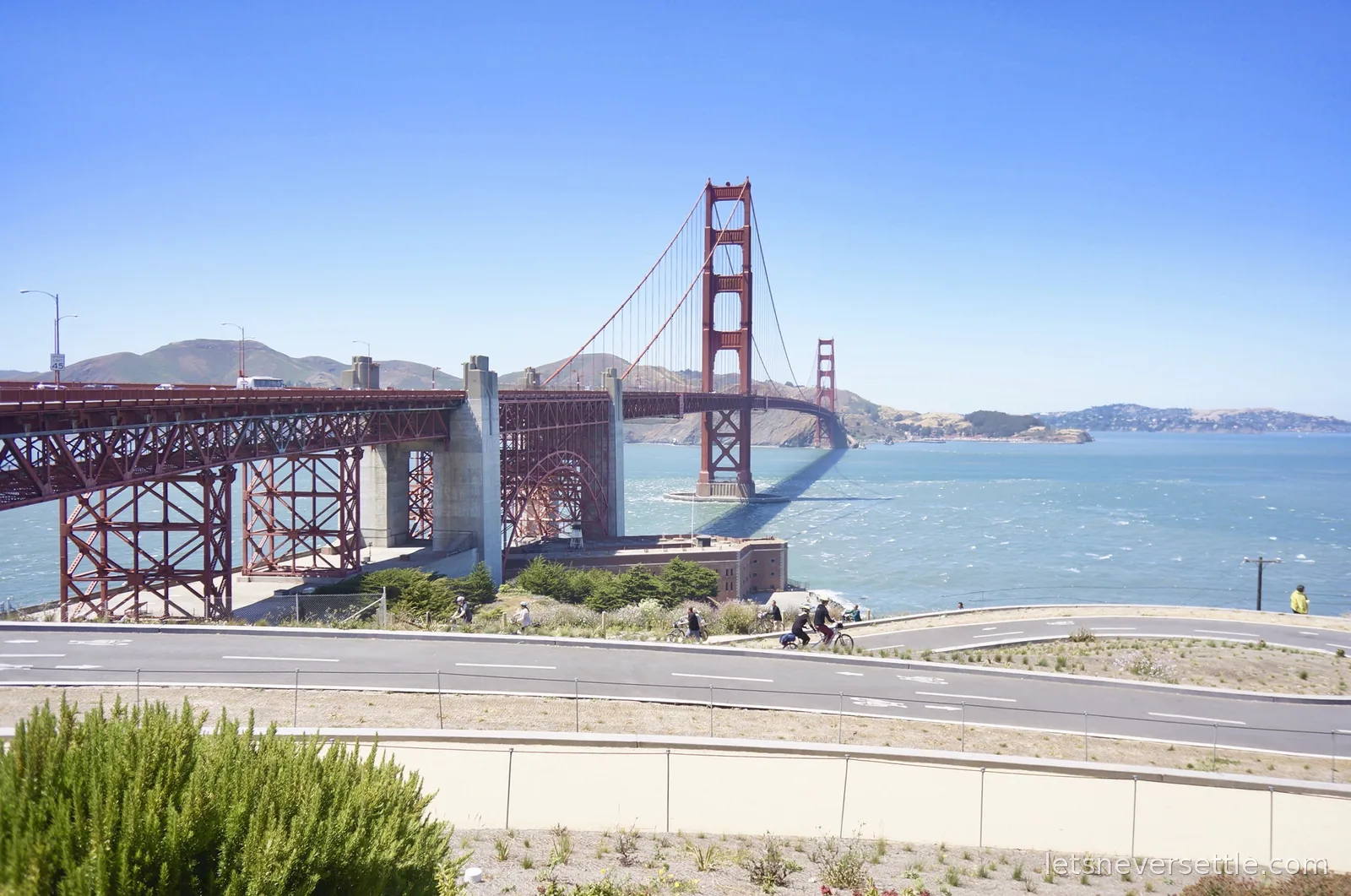 The Golden Gate Bridge spanning the strait with cyclists on the path below and the Marin Headlands behind