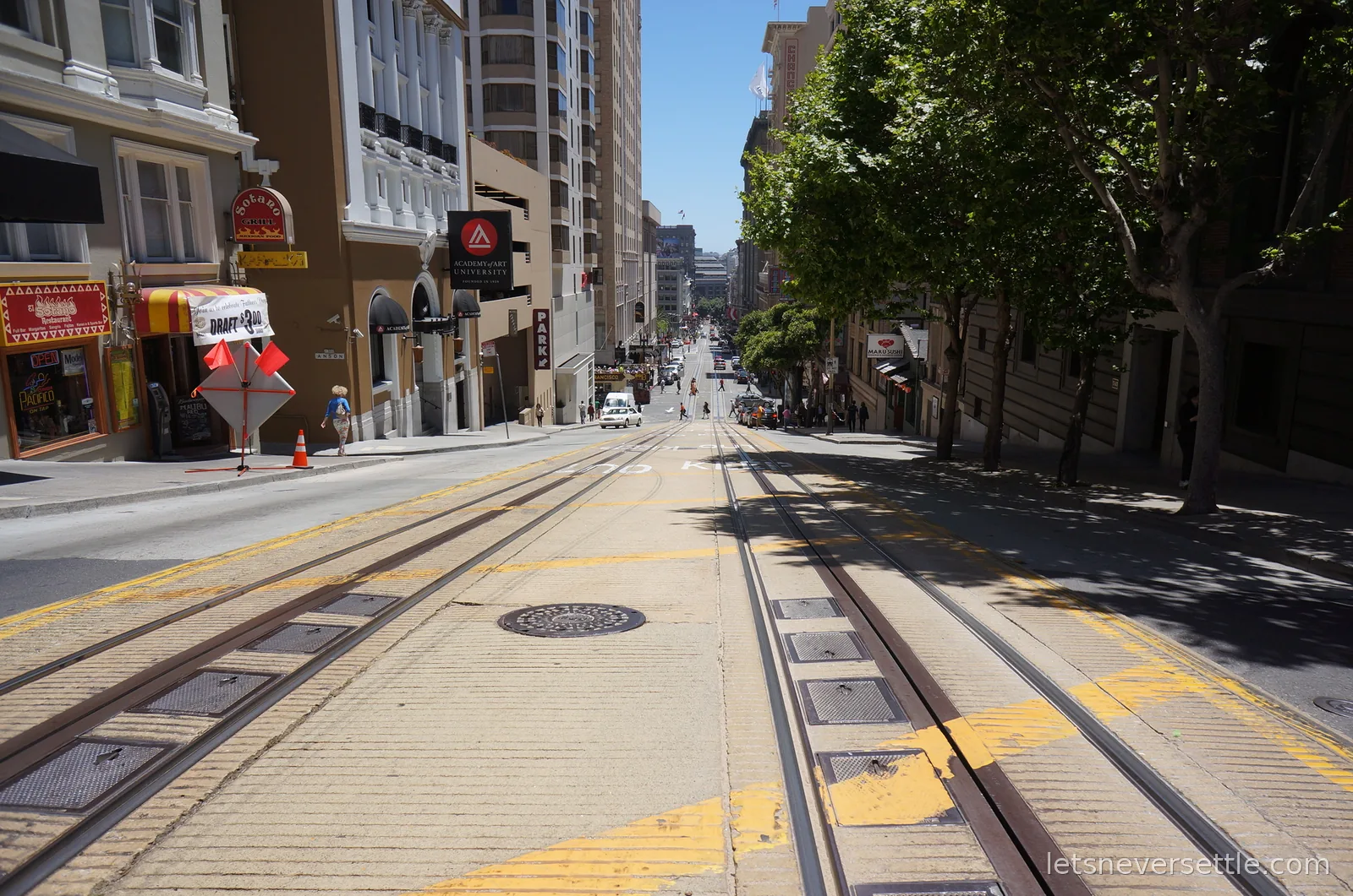Cable car tracks running down a steep San Francisco street between shops and trees towards downtown towers