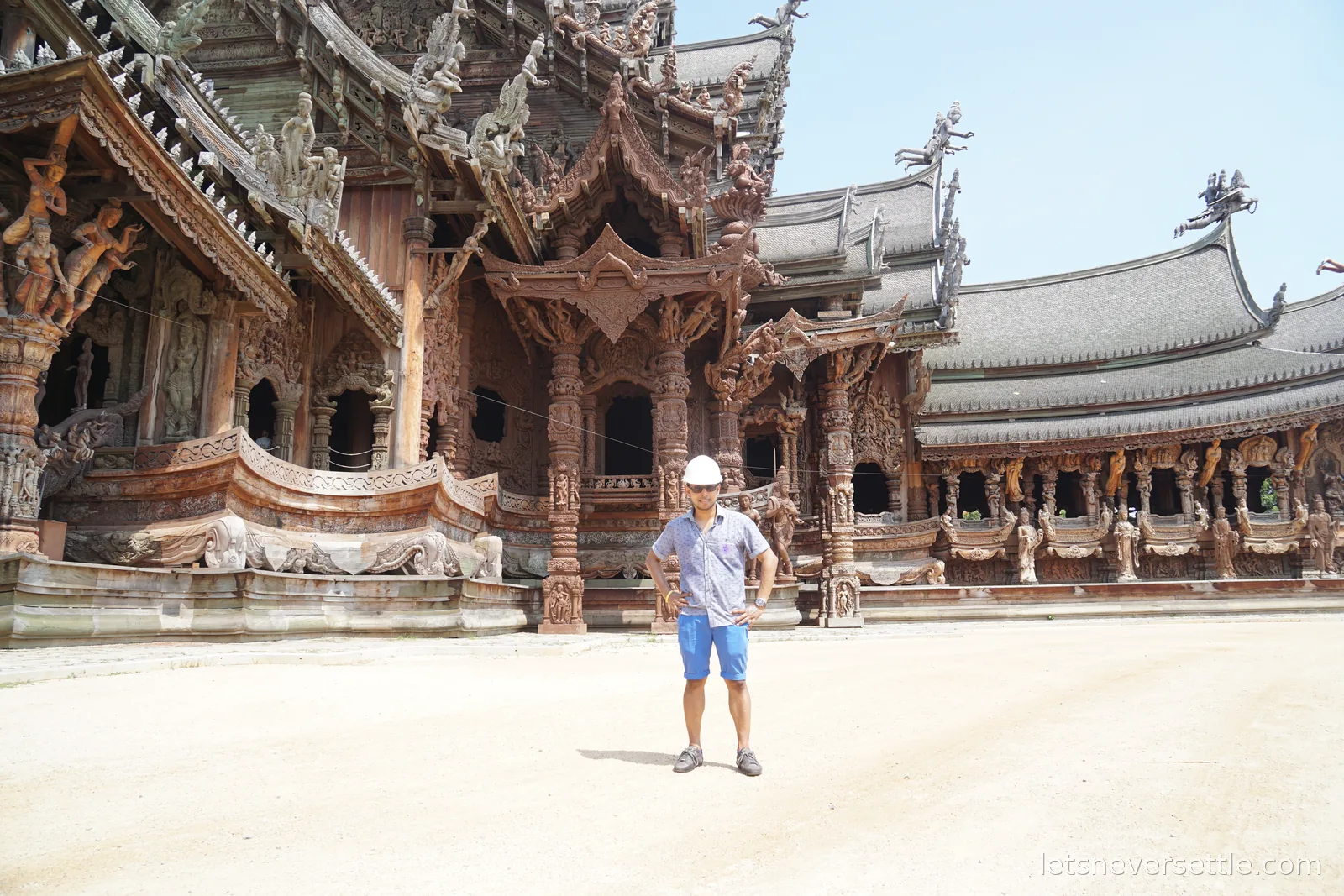 Dileep in a white hard hat standing in the empty sandy forecourt of the Sanctuary of Truth, in front of rows of carved wooden figures, pillars and porches