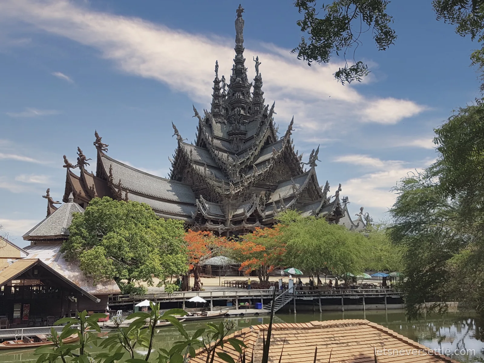 The Sanctuary of Truth seen across a lagoon, with bamboo rafts and long-tail boats moored below and a flame tree in orange bloom in front of it