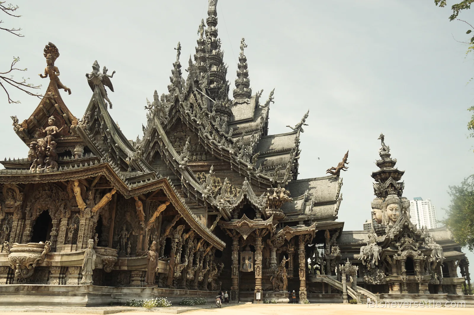 Wide view of the Sanctuary of Truth in Pattaya, a huge dark teak temple with layered gabled roofs and hundreds of carved figures, a modern high-rise visible behind it