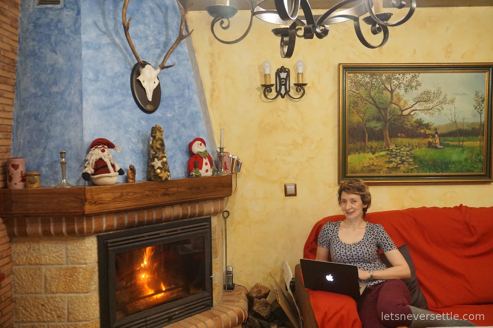 Inga sitting on a red sofa with a laptop beside a lit log burner, the mantelpiece decorated with Santa and snowman figures under mounted antlers