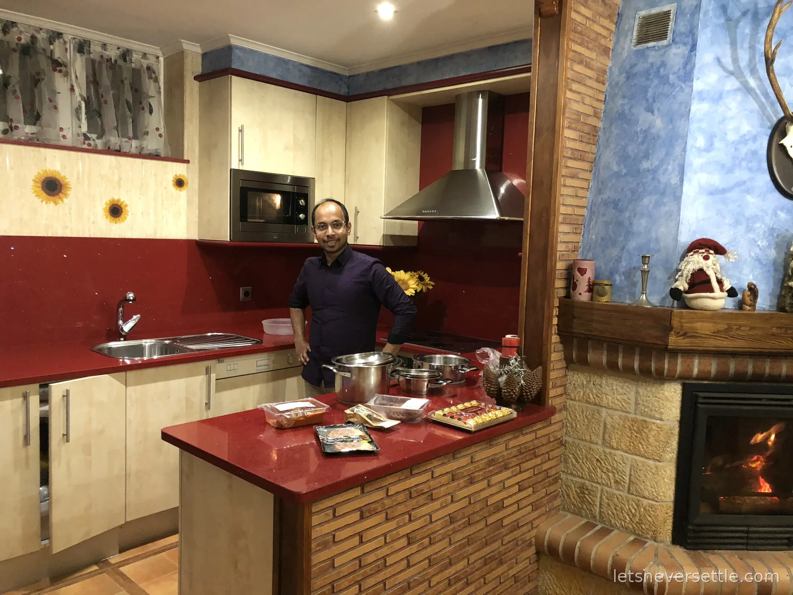 Dileep standing in a kitchen with a red sparkling counter, steel pots on the hob and packets of chorizo and a tray of chocolates laid out, a fire glowing to the right