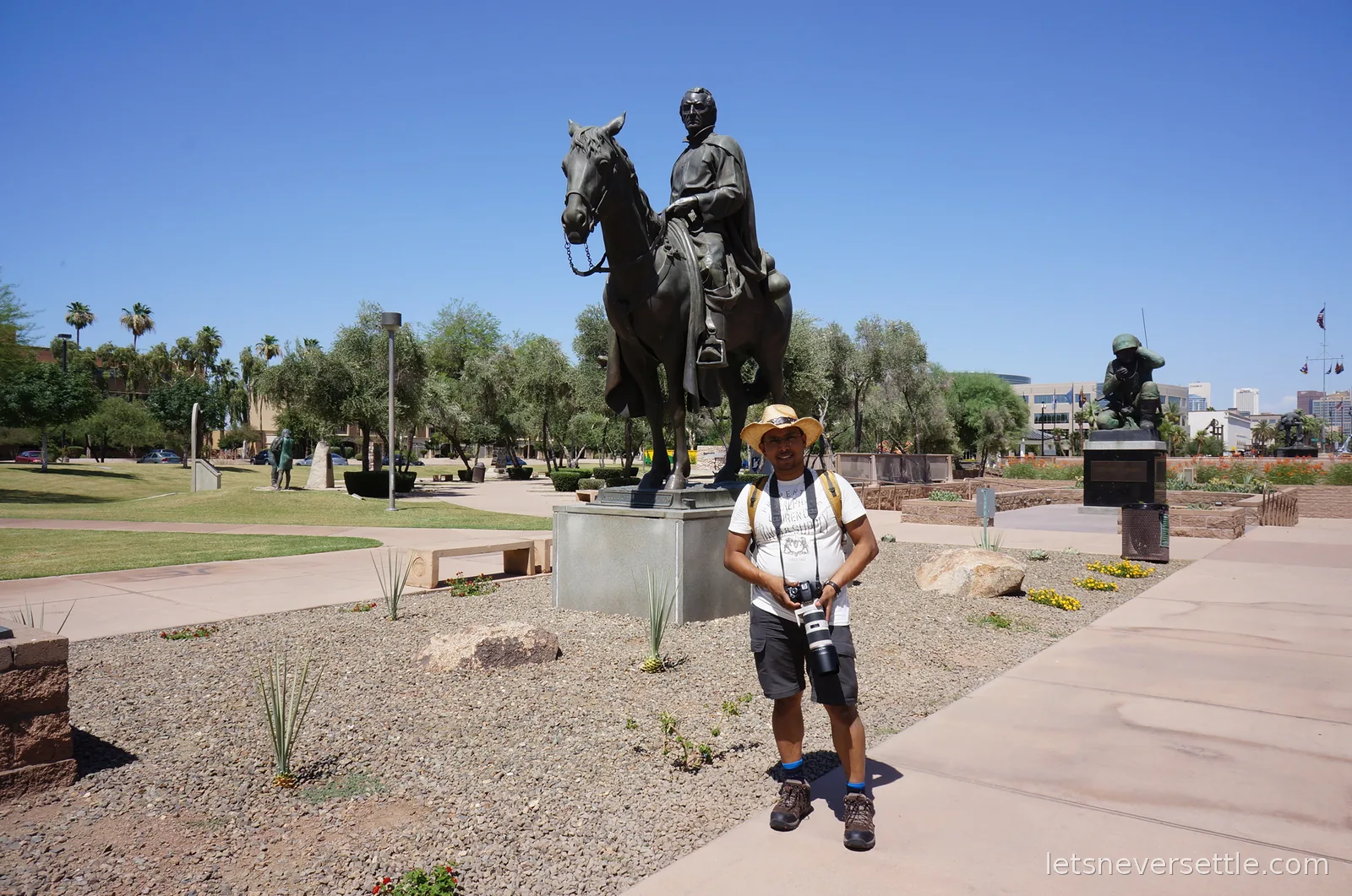 Dileep holding a long camera lens in front of a bronze equestrian statue on the memorial plaza by the Arizona State Capitol, Phoenix towers in the distance
