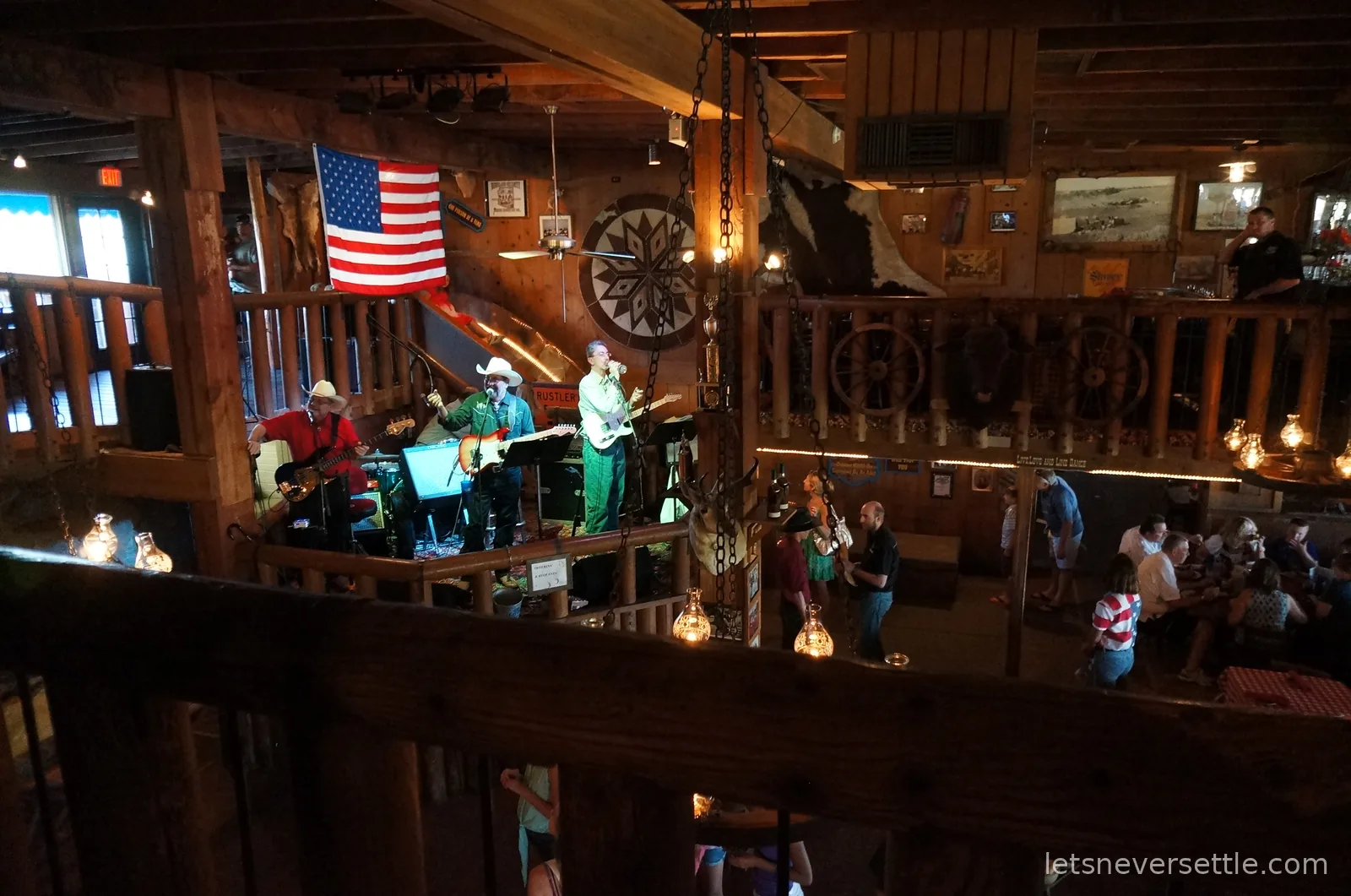 A country band in cowboy hats playing on a stage inside the timber dining hall, under a large American flag and a cowhide on the wall
