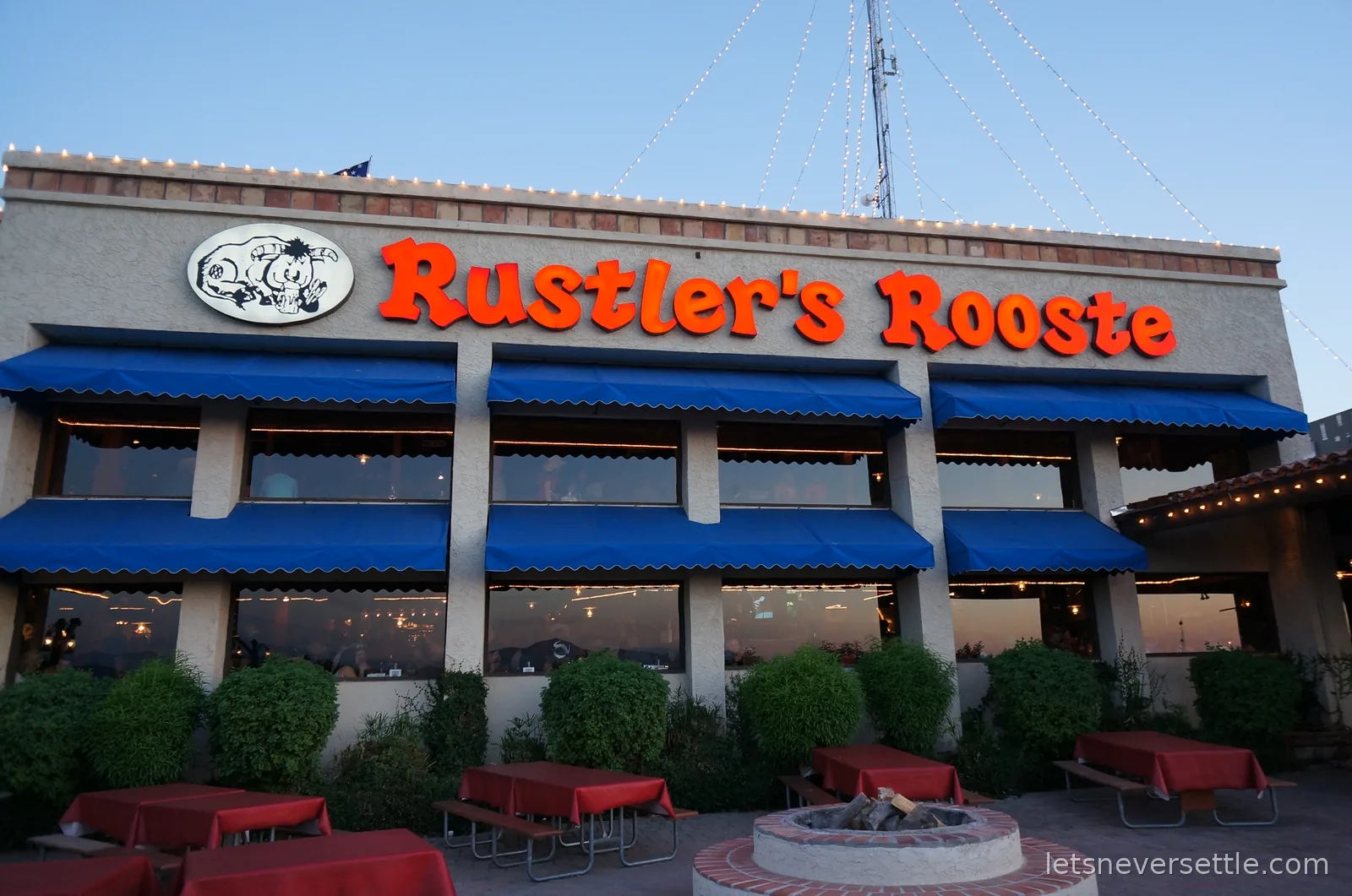 The Rustler's Rooste steakhouse exterior at dusk with blue awnings, red checked patio tables and a fire pit