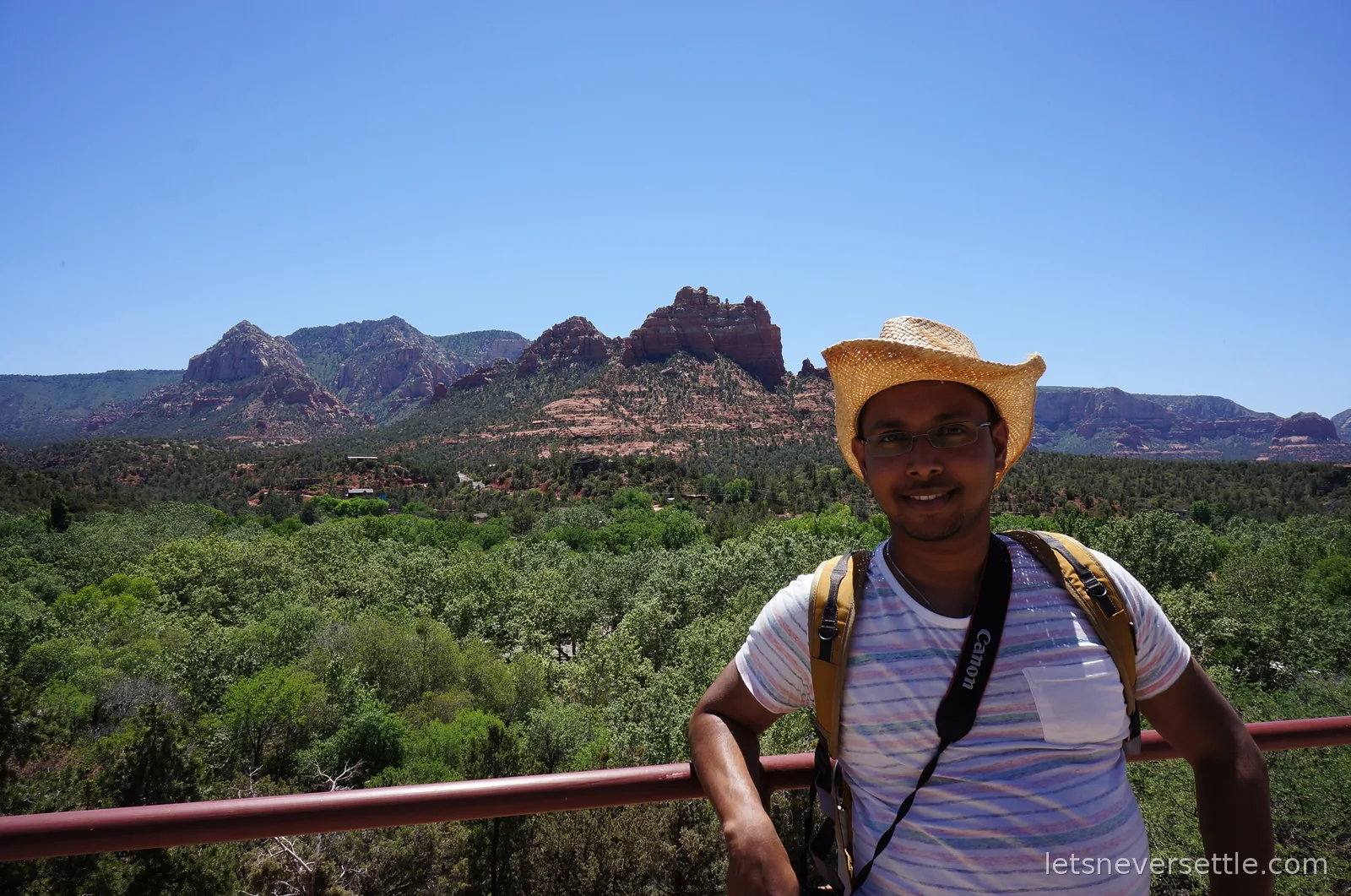 Dileep in a straw hat leaning on a red railing above the treetops, red rock formations across the valley