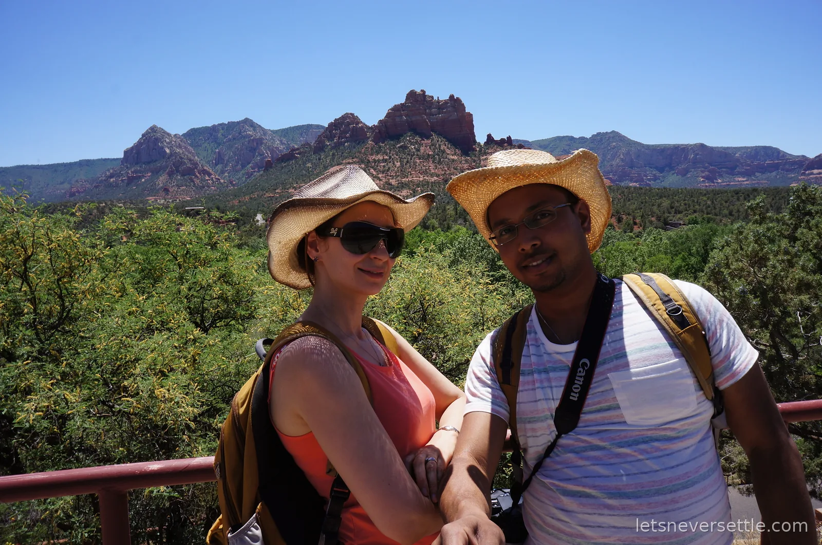 Selfie of Inga and Dileep in straw cowboy hats at a red-railed overlook with Sedona's red rock buttes rising beyond the trees