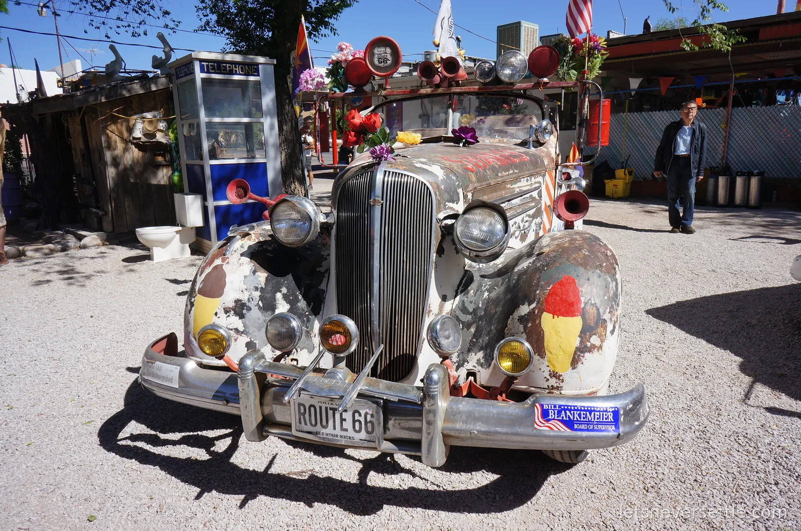 Front view of a rusted vintage car covered in horns, flowers and painted ice cream cones, with a Route 66 number plate