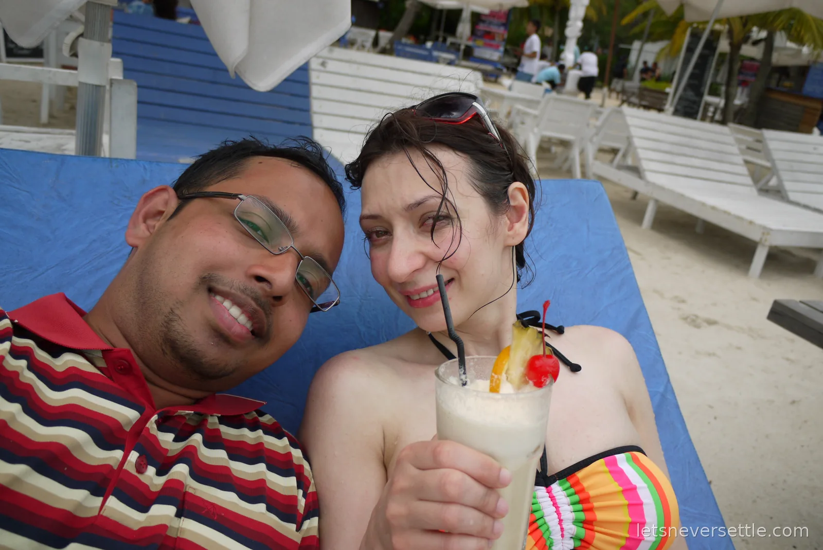 Inga and Dileep leaning together on a blue sun lounger with a pina colada dressed with pineapple and a cherry
